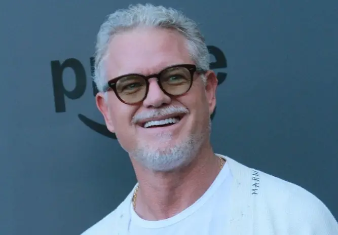 Eric Dane era um homem branco de cabelo e barba grisalhos. Na foto, ele usa blusa branca, cardigan da mesma cor e óculos de armação preta.
