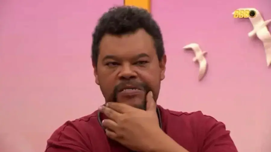 Homem pensativo com fundo rosa e decorativo de pombas sociais, uso de camisa vermelha, expressão facial de reflexão, ambiente de conversa ou entrevista.