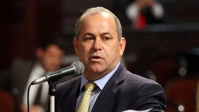 Homem sério em uma coletiva de imprensa, vestido com terno e gravata amarelo, falando em microfone em ambiente formal.