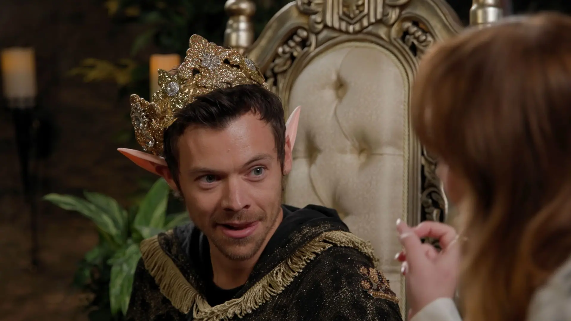 Harry Styles caracterizado como um monarca elfo, usando coroa e vestes elaboradas, sentado em um ambiente de fantasia com luz de velas.