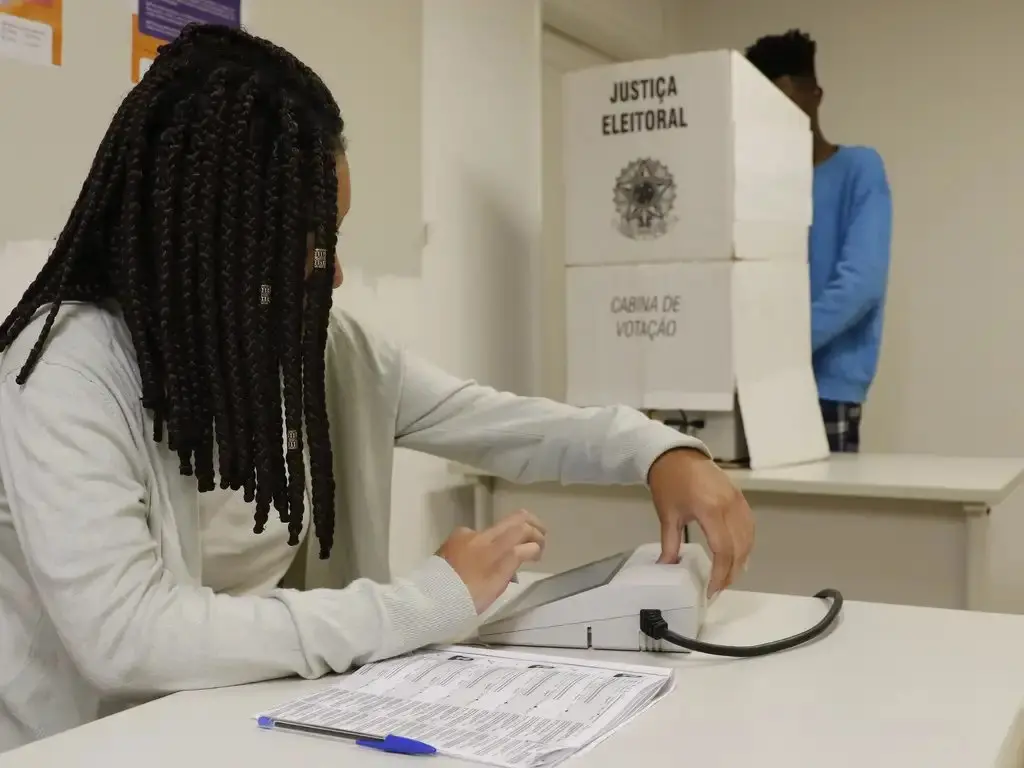 Mulher negra de tranças com roupa branca sentada em mesa de votação, com cabine de votação atrás.