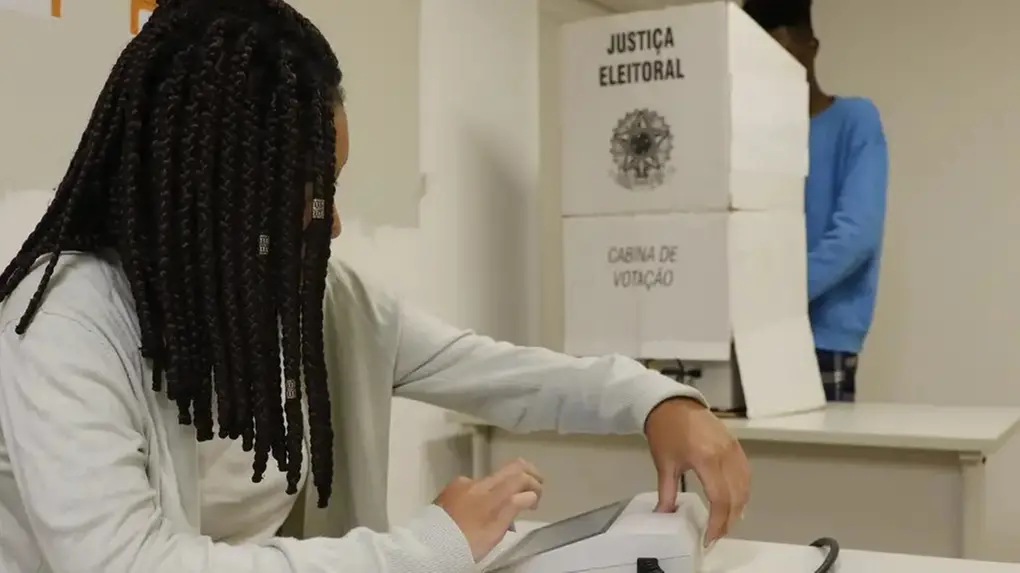 Mulher negra de tranças com roupa branca sentada em mesa de votação, com cabine de votação atrás.
