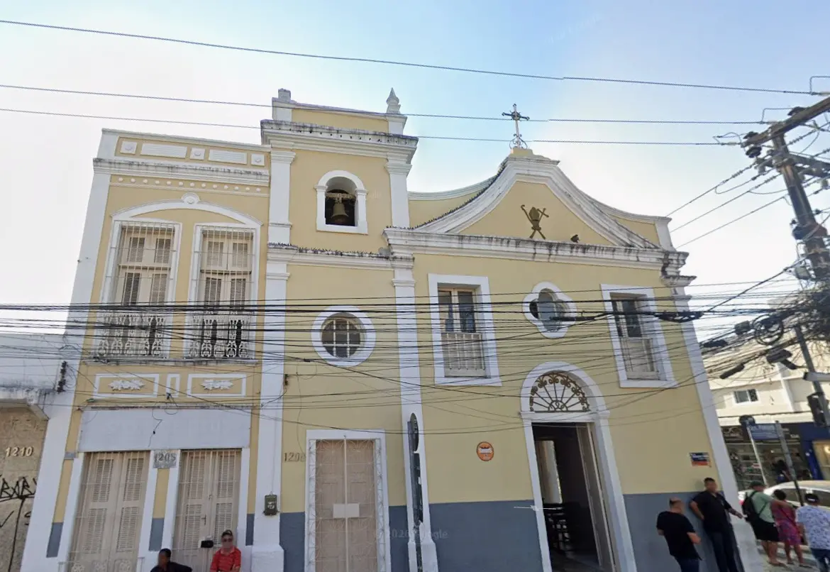 A fachada da Capela de São Bernardo apresenta tons de amarelo e detalhes brancos, destacando uma torre sineira com um sino de bronze sob um céu claro. A arquitetura colonial inclui janelas retangulares, uma entrada em arco e uma cruz no topo, emoldurada por uma rede de fios elétricos urbanos.