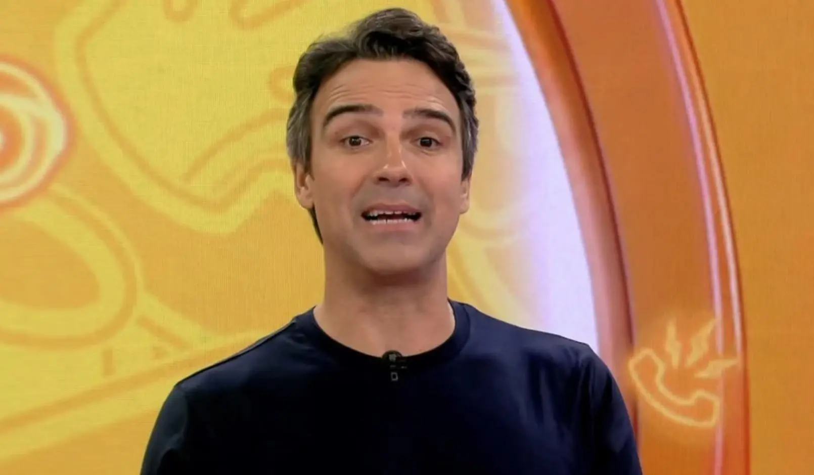 Pessoa em pé, vestindo uma camisa de manga longa escura, posicionada diante de um fundo colorido em tons de amarelo e laranja com elementos gráficos curvos.