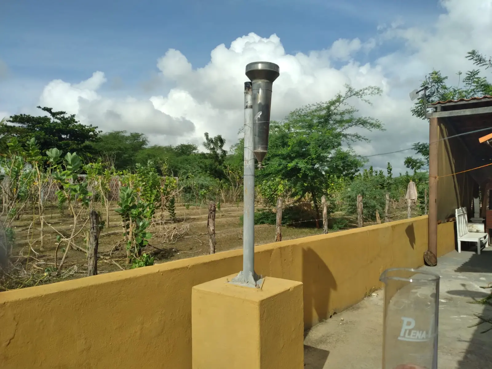 Imagem mostra um pluviômetro de matal em cima de um muro amarelo com uma plantação e o céu azul com nuvens ao fundo.