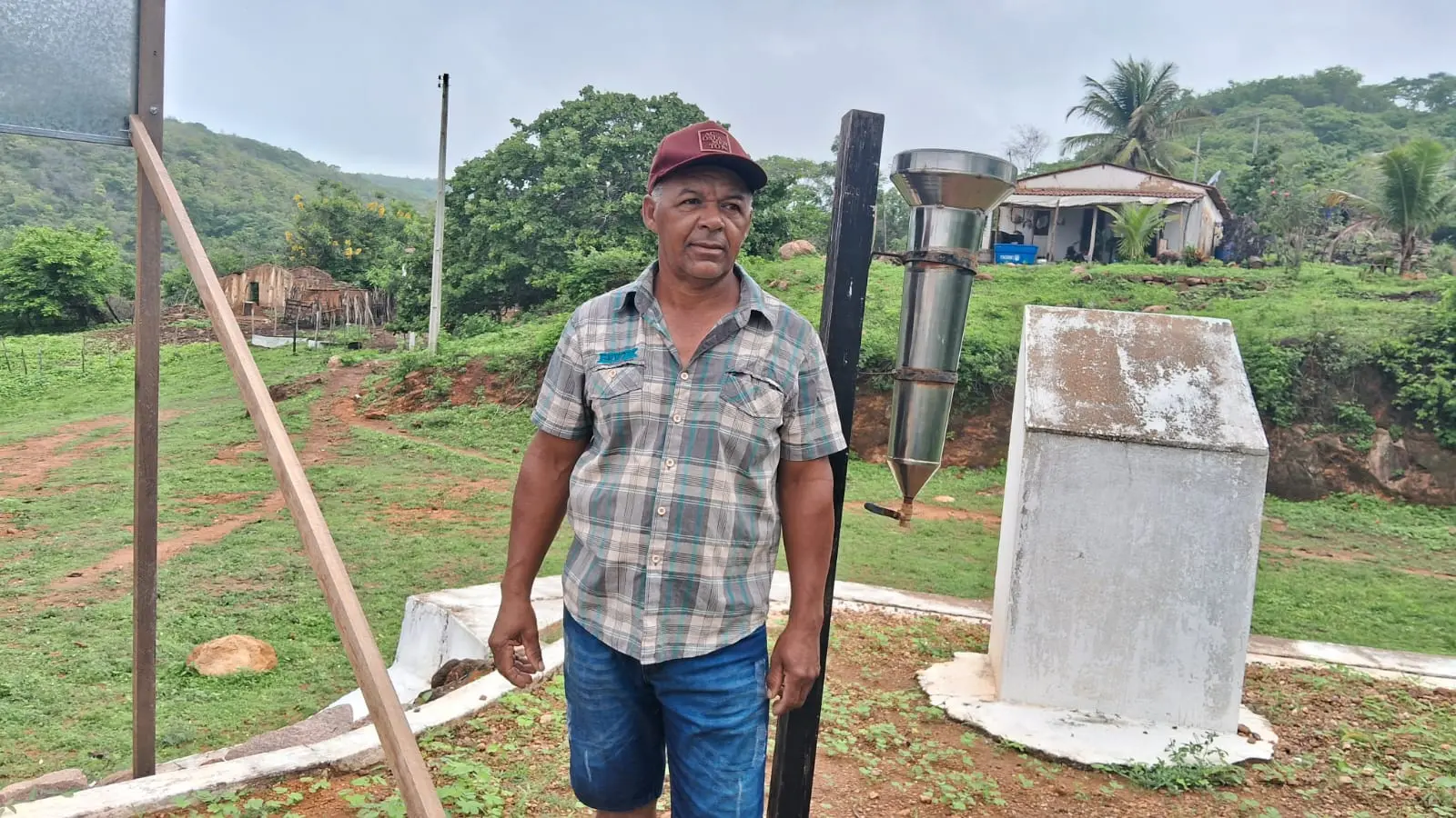 Imagem mostra José Roberto Tavares ao lado do pluviômetro pelo qual ele é responsável usando uma camisa xadrez, uma bermuda jeans azul e um boné vermelho.