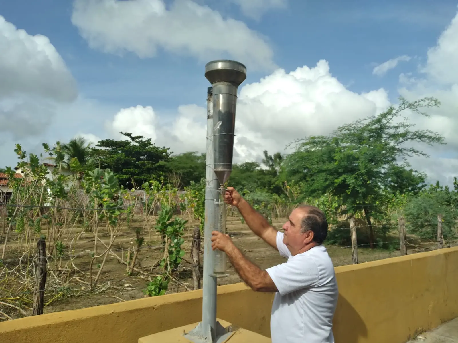 Imagem mostra Francisco das Chagas do Livramento ao lado do pluviômetro pelo qual é responsável em Limoeiro do Norte usando uma camiste branca, com uma plantação e o céu azul com nuvens ao fundo.
