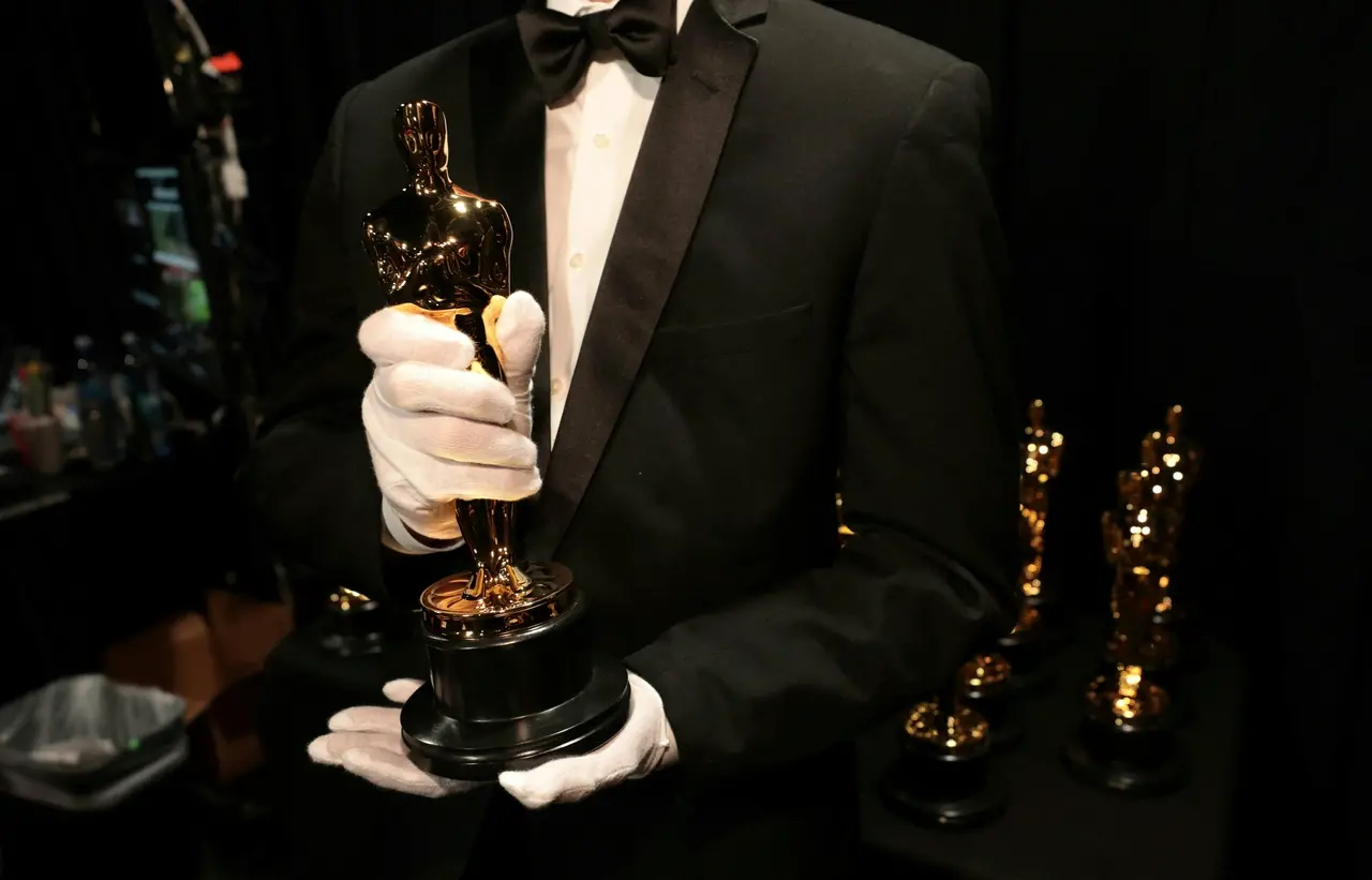 Uma pessoa vestindo um smoking clássico e luvas brancas segura cuidadosamente uma estatueta dourada do Oscar em primeiro plano. Ao fundo, outras estatuetas idênticas estão organizadas sobre uma superfície escura, criando uma composição elegante que destaca o brilho do prêmio contra o cenário sóbrio.