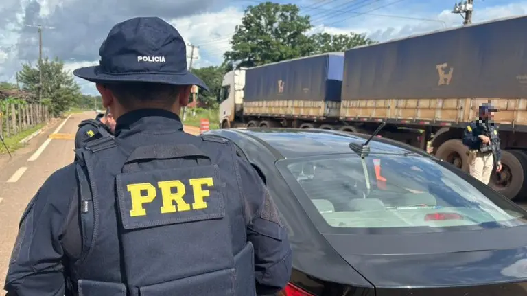 Agente da Polícia Rodoviária Federal sinaliza o trânsito durante fiscalização em rodovia, com veículos de carga e carro ao fundo.