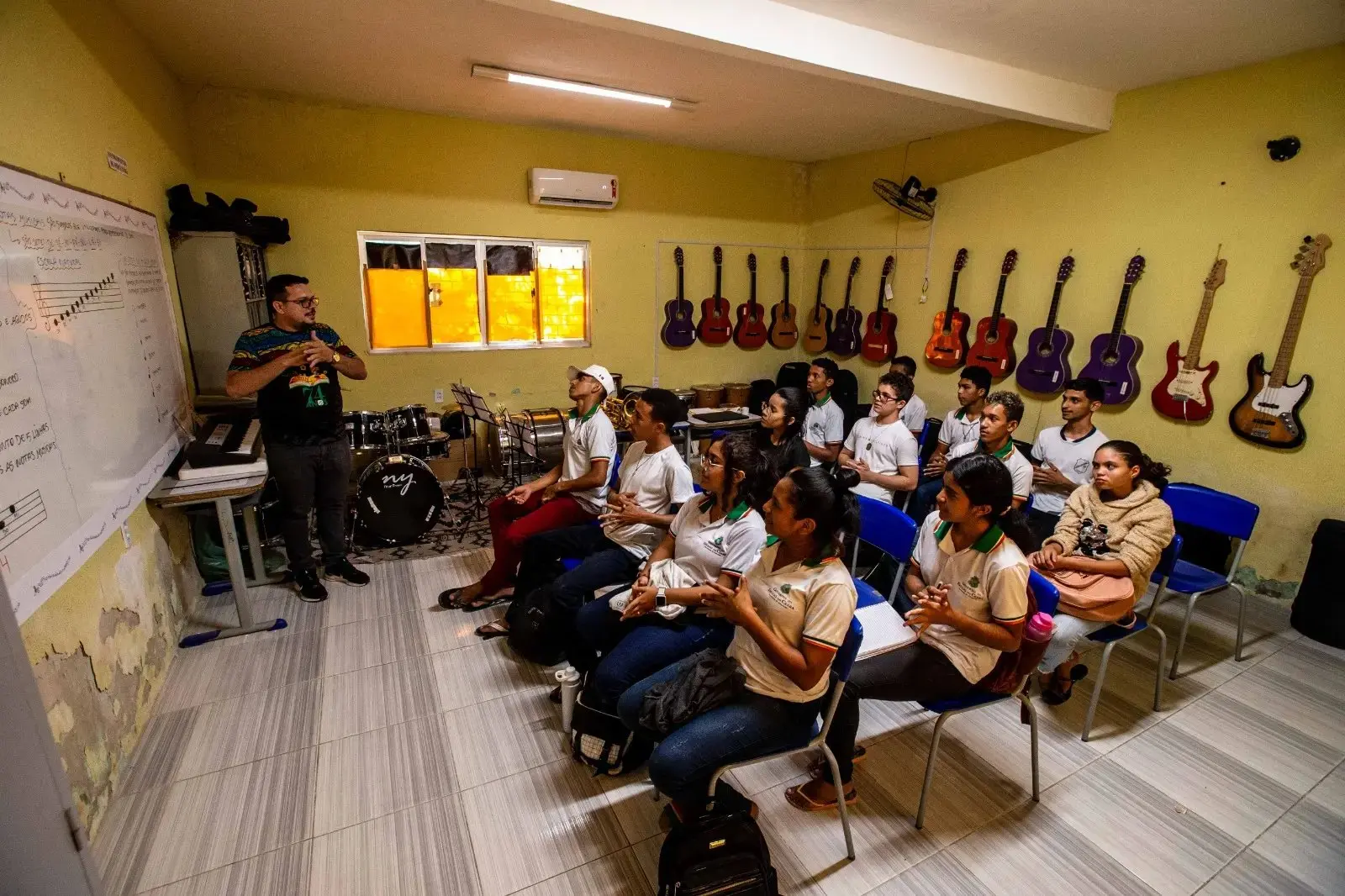 Sala de aula de música com cerca de 12 adolescentes sentados em cadeiras azuis, usando uniforme escolar, enquanto um professor em pé conduz a atividade na frente do grupo. Nas paredes, há vários violões pendurados e, ao fundo, uma bateria e outros instrumentos musicais, indicando um ambiente de ensino artístico em tempo integral.