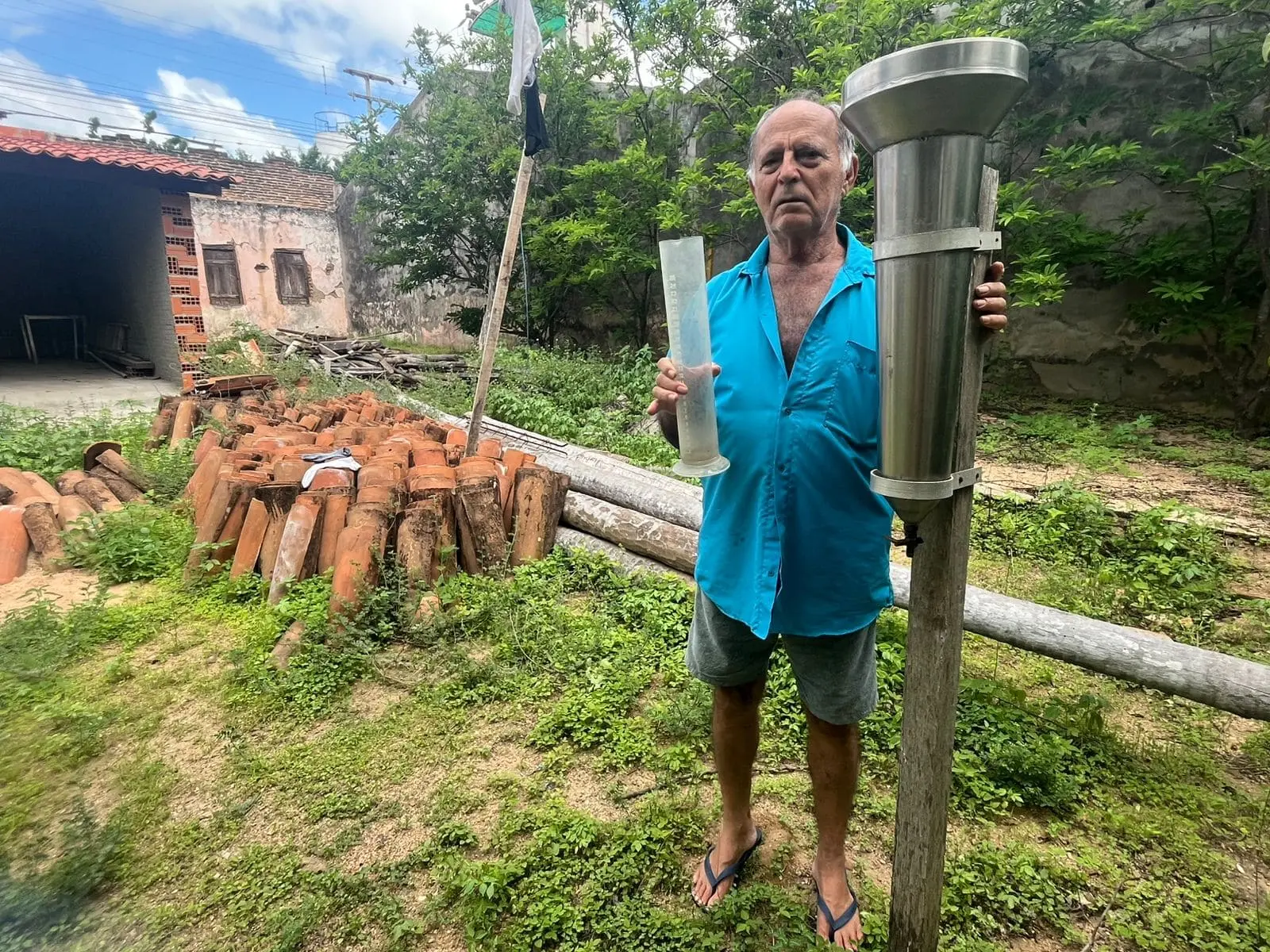 Imagem mostra José Alencar ao lado do pluviômetro pelo qual ele é responsável usando uma camisa de botão azul e uma bermuda.