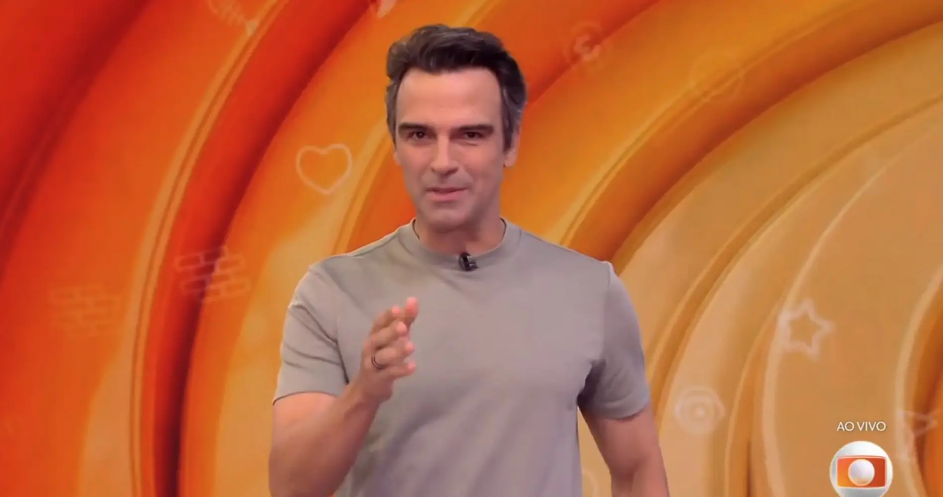 Homem apresentador de televisão sorridente em um cenário colorido de fundo laranja, com detalhes decorativos. Cena de programa de TV ao vivo.