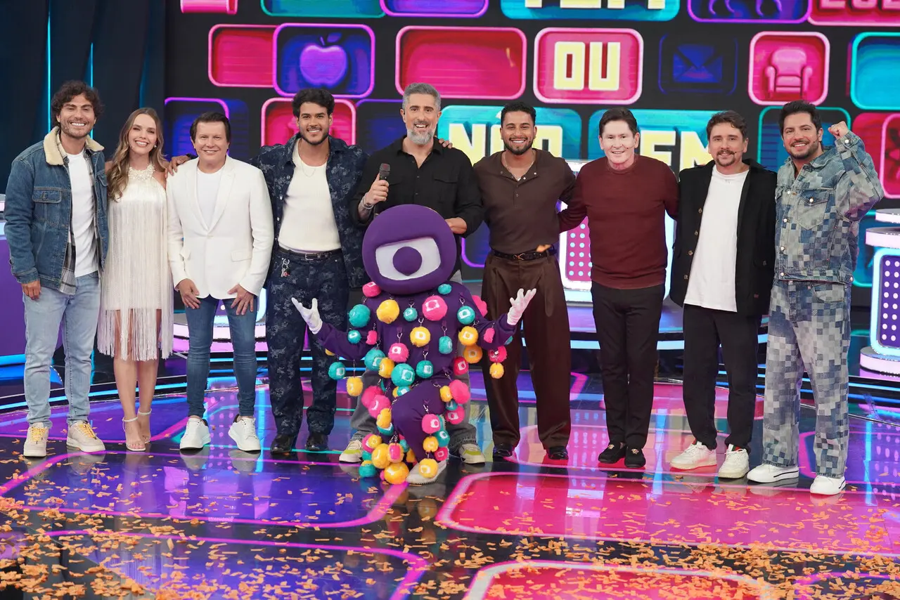 Um grupo de nove pessoas está posicionado lado a lado sobre um palco colorido com luzes brilhantes e painéis eletrônicos ao fundo exibindo formas geométricas e ícones. À frente do grupo, há um mascote roxo com um grande olho central e um traje coberto por esferas multicoloridas. O chão do palco está decorado com confetes laranja espalhados, e o cenário tem um visual vibrante e festivo.