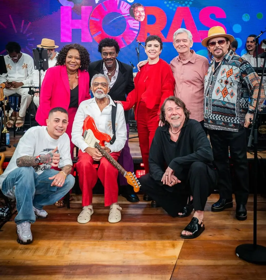 Grupo de pessoas reunidas em um palco iluminado, posando para uma foto em frente a um telão colorido com a palavra ‘HORAS’. No centro, uma pessoa sentada segura uma guitarra elétrica vermelha e branca. Outras pessoas estão em pé ou agachadas ao redor, vestindo roupas variadas e coloridas, incluindo ternos vibrantes, peças estampadas e acessórios como chapéus. Ao fundo, músicos e instrumentos aparecem no cenário, sugerindo um ambiente de apresentação musical.