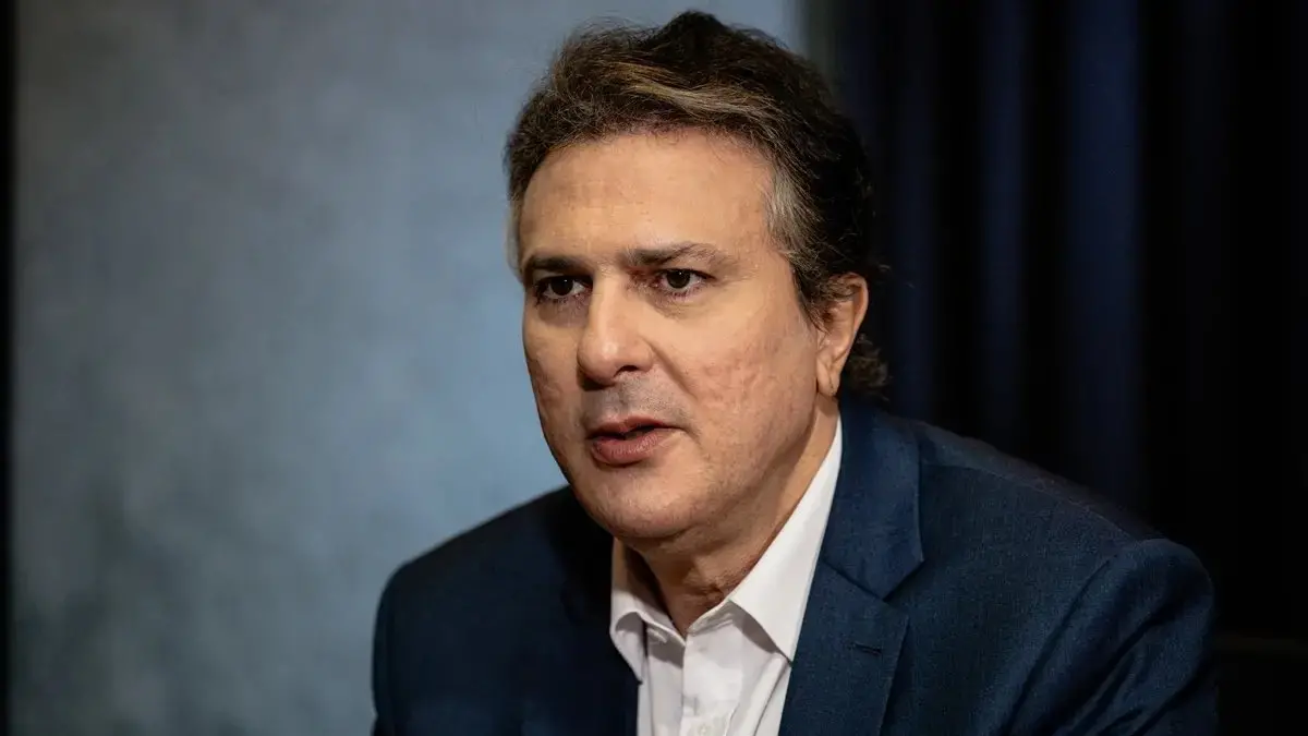 Fotografia em close-up do Ministro da Educação, Camilo Santana. Ele é um homem de pele clara, cabelos castanhos levemente grisalhos e usa um paletó azul-marinho sobre uma camisa social branca. Camilo está posicionado de perfil parcial, com uma expressão séria e lábios entreabertos, durante uma entrevista. O fundo está desfocado em tons de cinza e azul escuro.