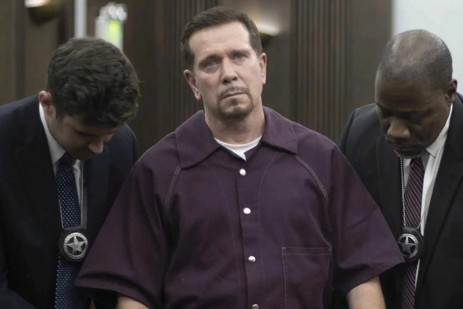 Imagem do ator bobby j brown, como um personagem sendo escoltado por policiais durante um procedimento policial, com expressão séria e roupas de prisão.