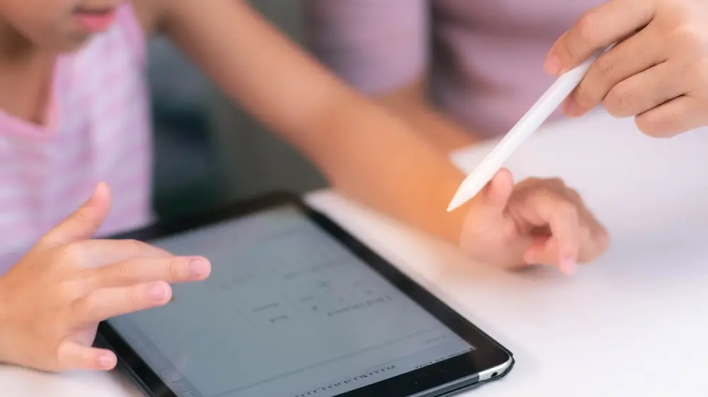 Na imagem, fotografia em close-up focada nas mãos de uma criança e de uma pessoa adulta sobre uma mesa branca. A criança interage com a tela de um tablet, enquanto a pessoa adulta segura uma caneta digital branca, apontando em direção ao dispositivo. A cena sugere um momento de aprendizado mediado por tecnologia.
