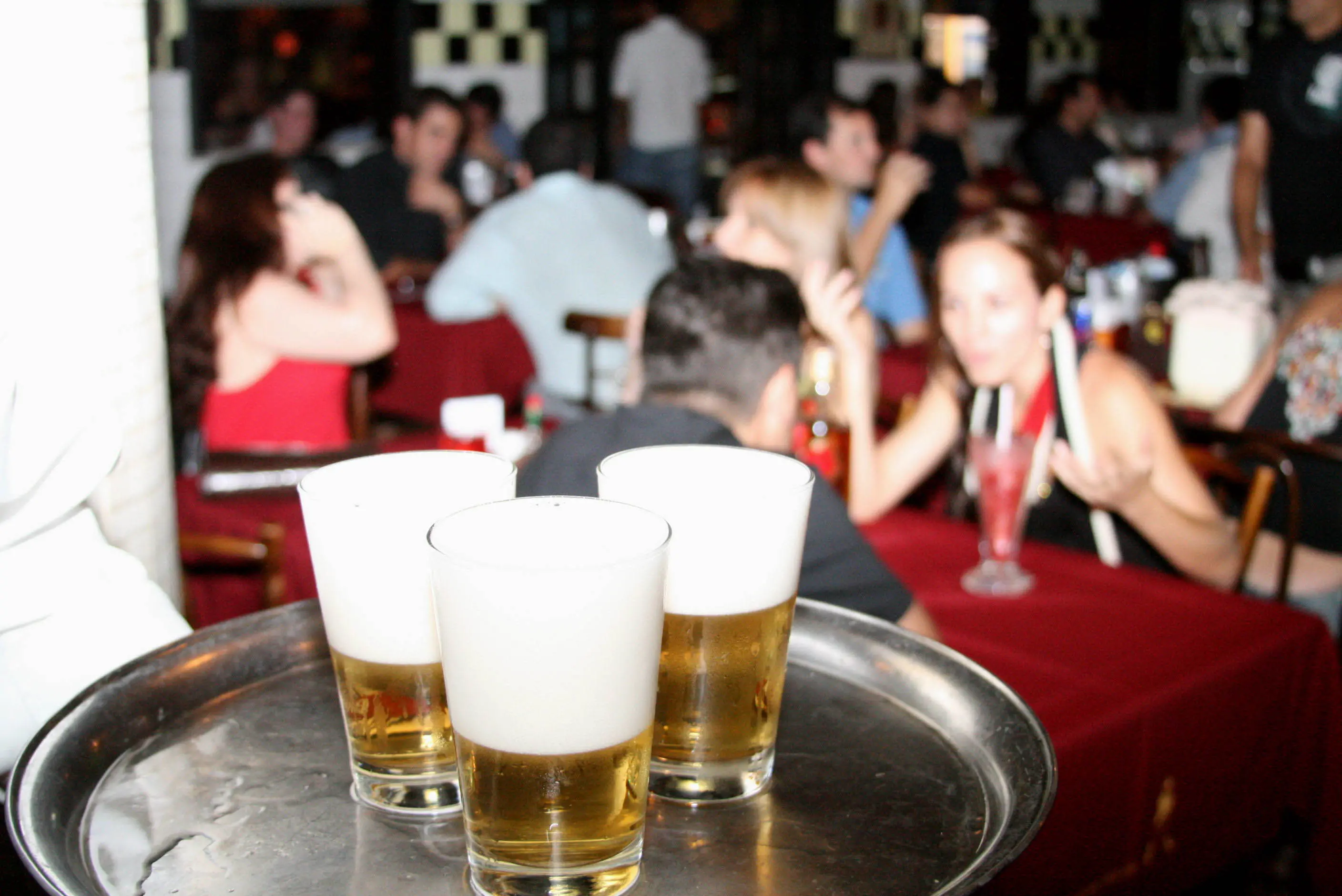 Garçom carregando uma bandeja com três copos de cerveja, com um restaurante ou bar movimentado desfocado ao fundo.