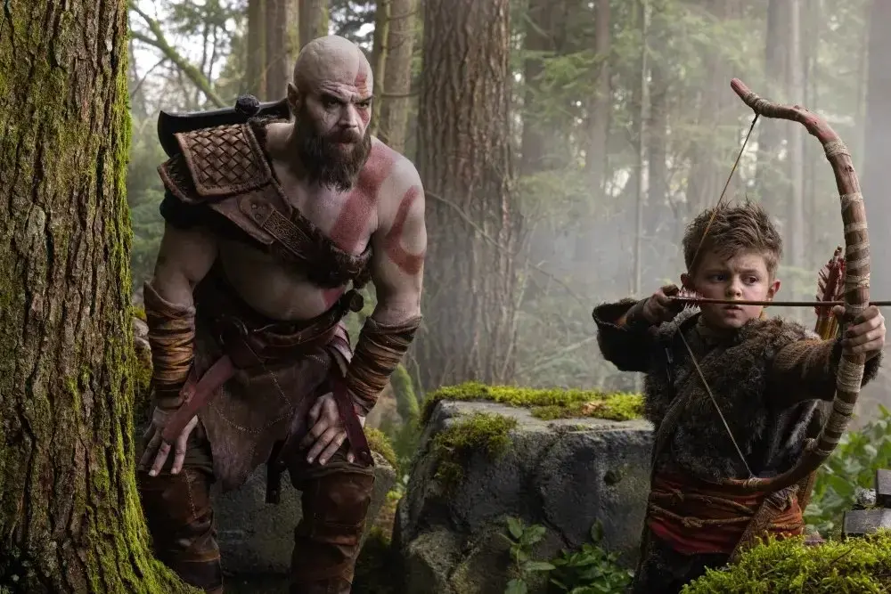 Kratos, interpretado por Ryan Hurst no live action, observa atentamente enquanto o jovem Atreus aponta seu arco e flecha em uma floresta densa e ensolarada na primeira imagem de divulgação da série God of War, baseada nos jogos. A dupla veste trajes de couro e peles detalhados, compondo uma cena de treinamento vibrante que destaca a tensão e o foco entre pai e filho.