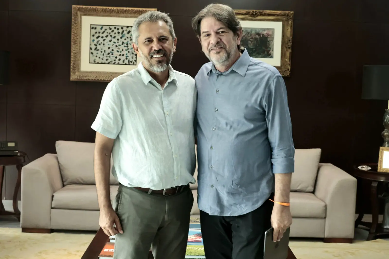 O governador do Ceará, Elmano de Freitas, à esquerda, e o senador Cid Gomes, à direita, posam sorridentes lado a lado em um ambiente institucional. Elmano veste uma camisa de mangas curtas clara e calça social, enquanto Cid Gomes usa uma camisa social azul de mangas compridas e segura um tablet. Ao fundo, vê-se um sofá cinza e quadros emoldurados na parede.