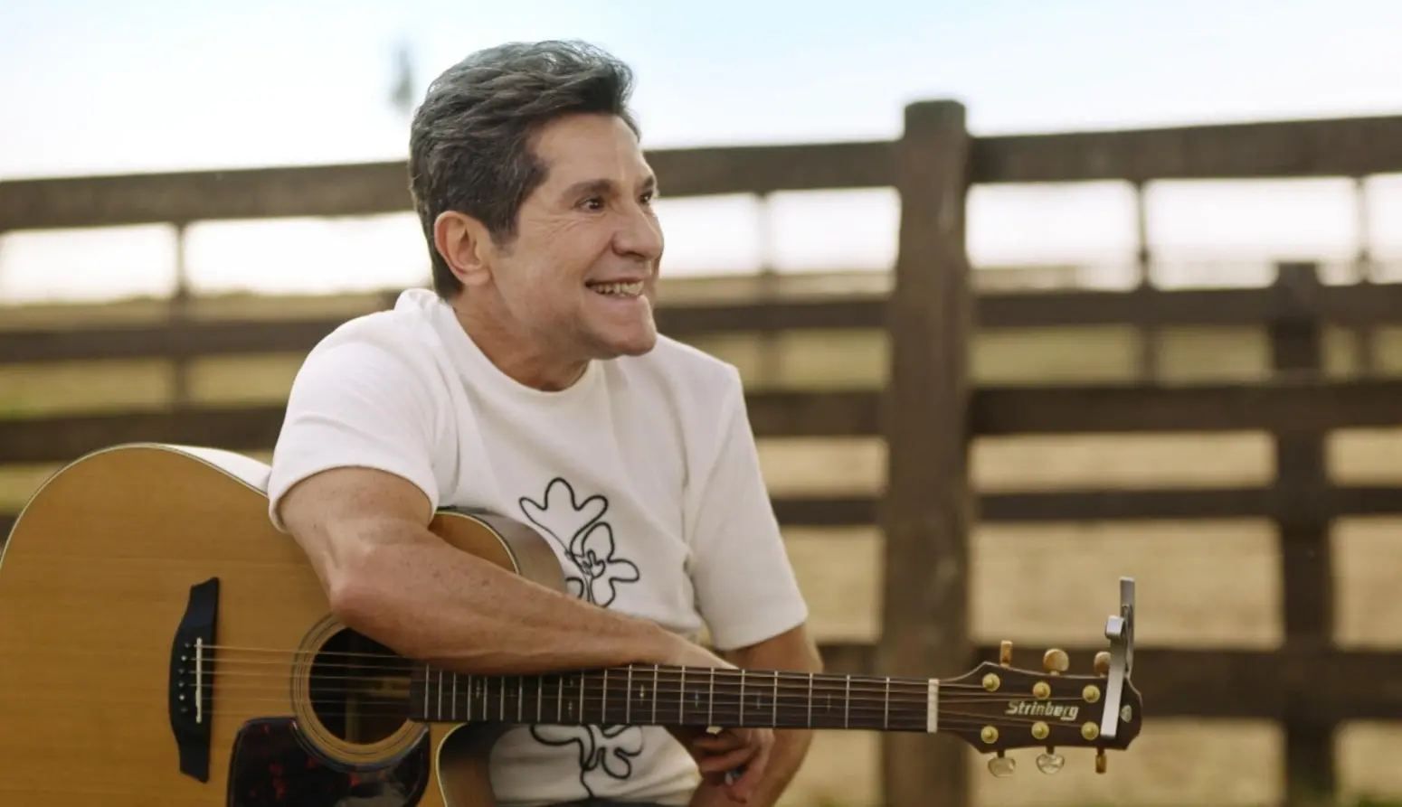 Daniel é um homem branco de meia idade e cabelo preto. Na foto, ele está sentado em uma fazenda com um violão sobre o colo.