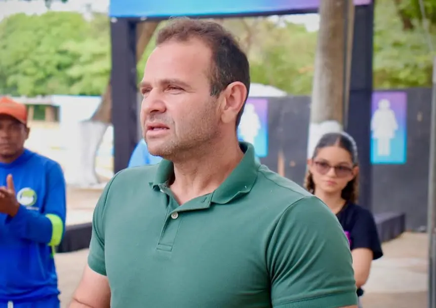 Fotografia em plano médio do ex-prefeito de Aracoiaba, Edim Oliveira. Ele é um homem de pele clara, cabelos curtos e castanhos, e veste uma camisa polo verde escura. Ele olha para o lado com uma expressão séria. Ao fundo, de forma levemente desfocada, aparecem outras pessoas e estruturas de um evento ao ar livre.