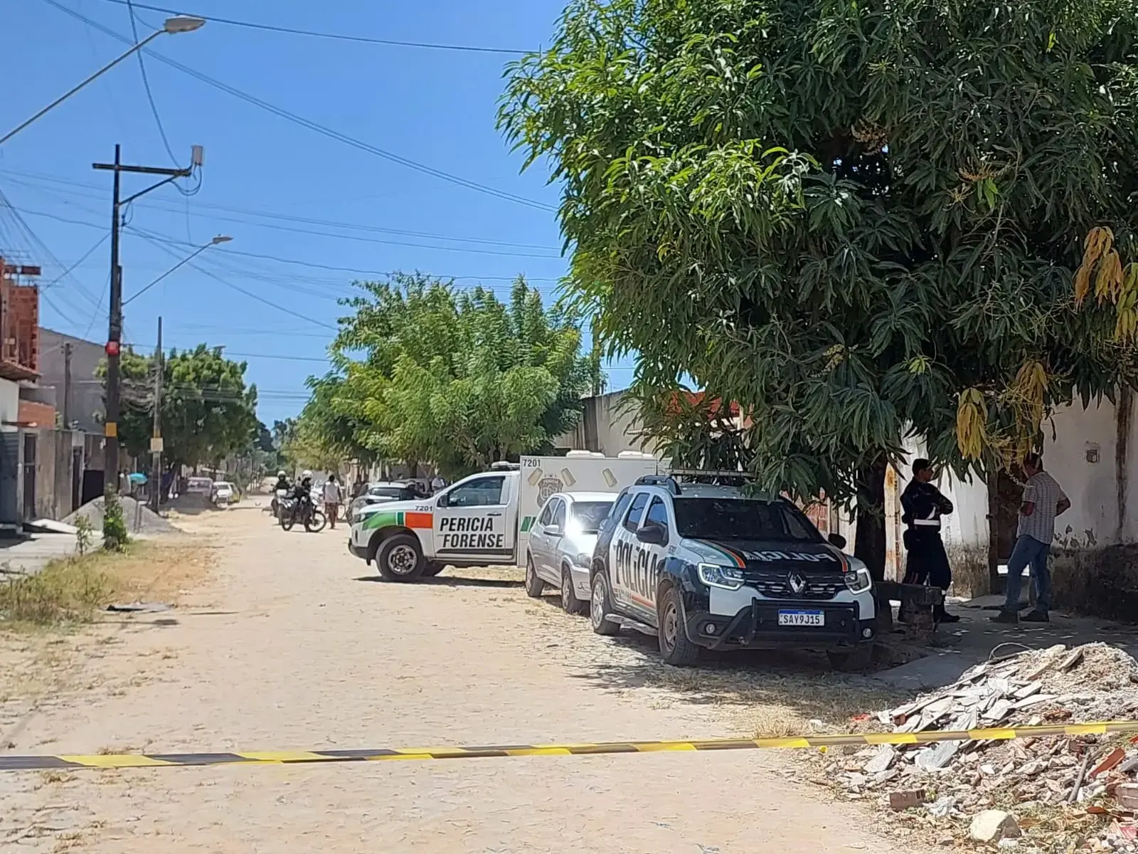 Rua de terra isolada por fita policial com viaturas da Polícia Militar e da Perícia Forense estacionadas próximo a uma residência, em área residencial com árvores e casas simples, durante atendimento a ocorrência criminal.