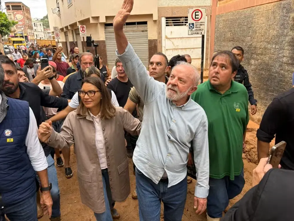 Na imagem, o presidente Luiz Inácio Lula da Silva e a primeira-dama Janja Lula da Silva caminham por uma rua estreita e íngreme, coberta de lama, em Minas Gerais. Lula, ao centro, veste camisa azul clara e calça jeans, e está com o braço direito erguido, apontando para algo acima ou acenando. Janja caminha ao seu lado esquerdo, de óculos, camisa clara e um casaco bege. Eles estão cercados por uma multidão de apoiadores, seguranças e moradores que registram o momento com celulares. À direita, um homem de camisa verde observa atentamente a direção para onde Lula aponta. O cenário ao fundo mostra construções simples, uma placa de trânsito de proibido estacionar e marcas de barro nas paredes, evidenciando os danos causados por chuvas ou deslizamentos na região.