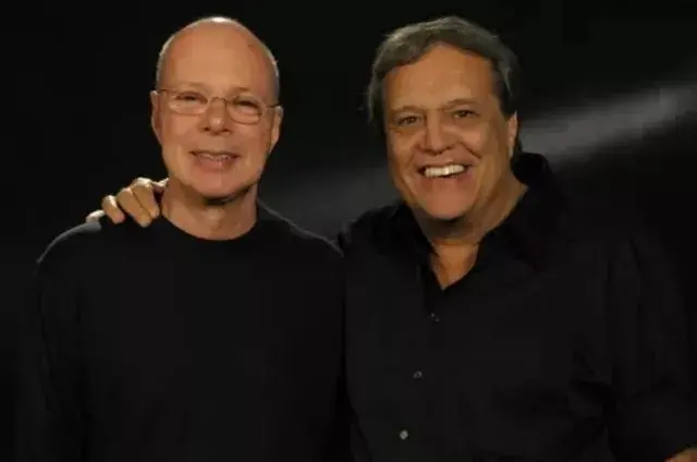 Na imagem, fotografia de plano médio de dois homens sorridentes, Gilberto Braga e Dennis Carvalho, posando abraçados contra um fundo preto. À esquerda, Gilberto é careca, usa óculos de grau discretos e uma camisa preta de manga longa. À direita, Dennis tem cabelos grisalhos e veste uma camisa social preta de botões. A iluminação é suave, destacando o rosto de ambos.