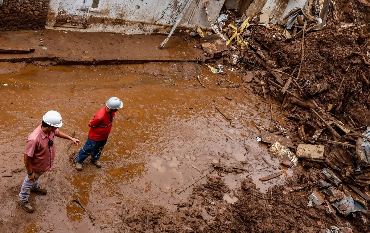 Dois trabalhadores com capacetes de proteção caminham por área tomada por lama após destruição em cenário de desastre ambiental.A foto mostra a busca por desaparecidos após fortes chuvas em Juiz de Fora e Ubá.