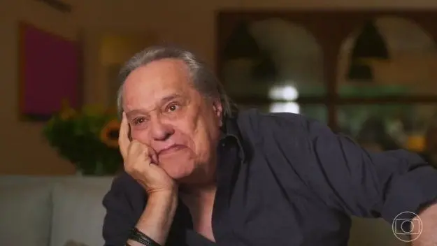 Imagem mostra o diretor Denis Carvalho com a mão no rosto e mo dedo na fonte direita em uma pose pensativa, de camisa escura.