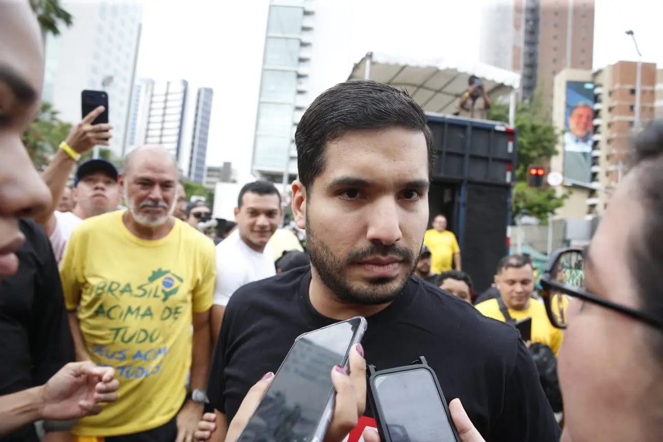 Homem de barba e camisa preta é cercado por pessoas e celulares durante entrevista em meio a uma multidão em área urbana; ao fundo, apoiadores vestem camisetas amarelas com a frase “Brasil acima de tudo, Deus acima de todos”, e há prédios altos e um trio elétrico na rua.