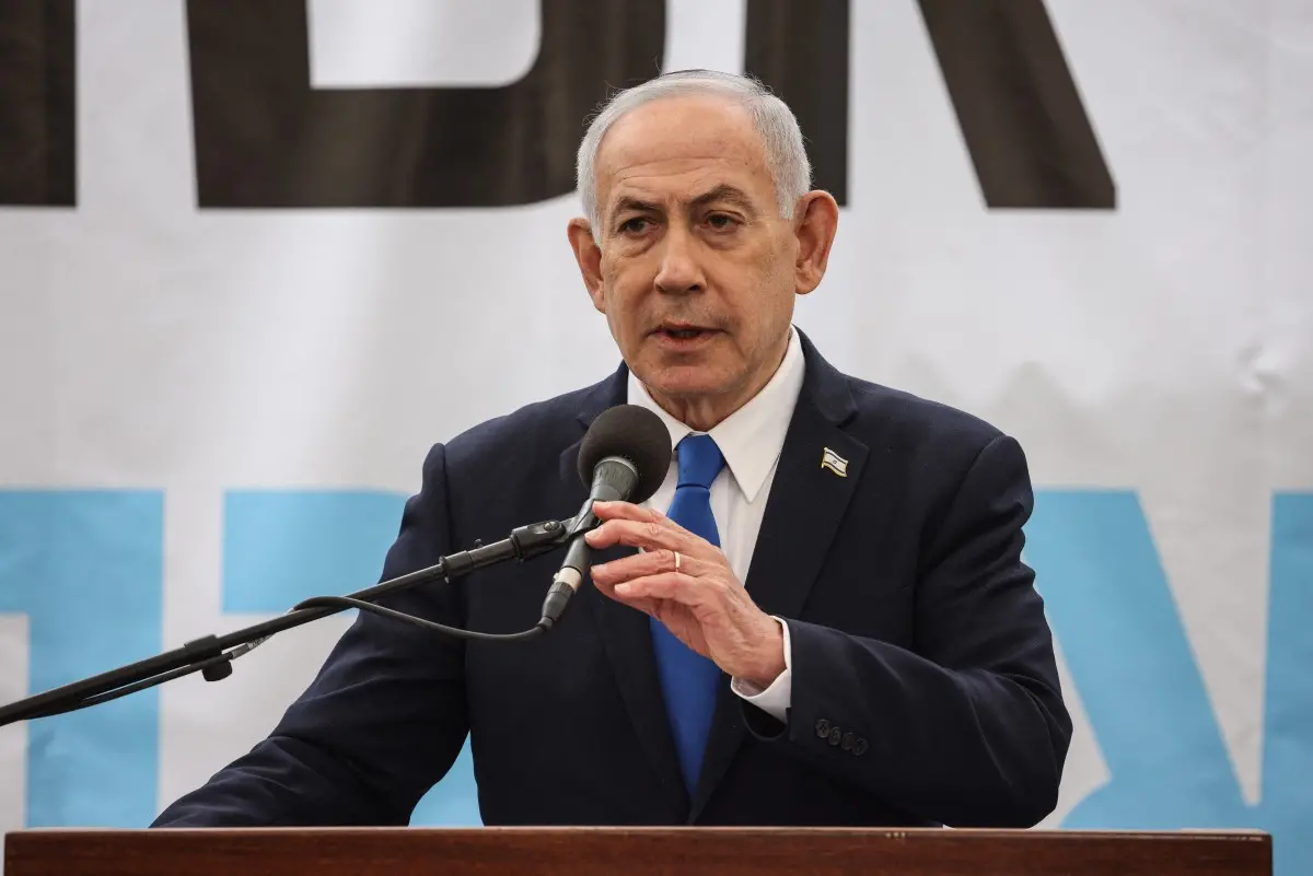 Benjamin Netanyahu, primeiro-ministro de Israel, falando em um evento com fundo branco e uma bancada de madeira, usando terno e gravata azul.