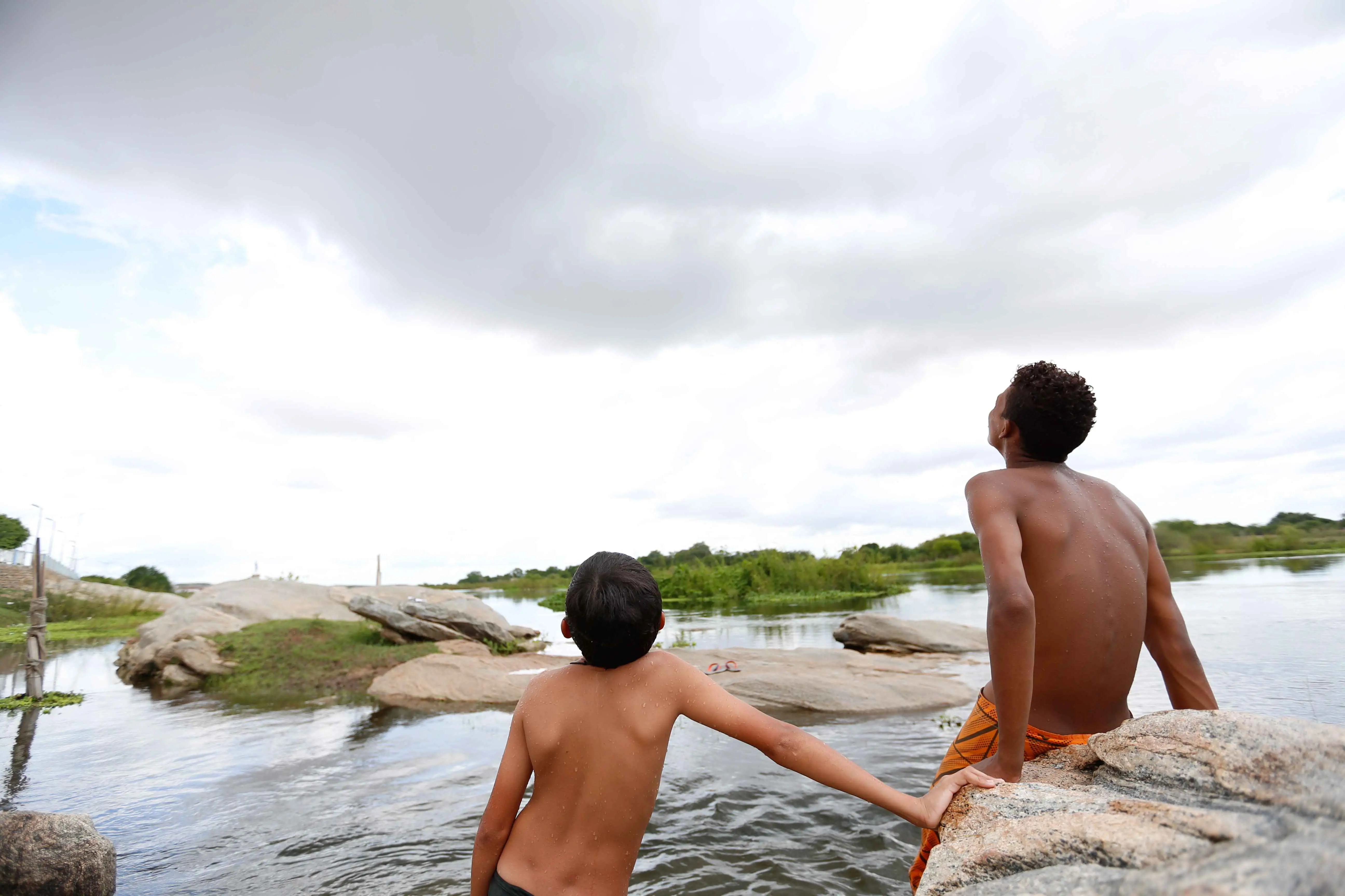 Dois meninos à beira de um rio, um sentado em uma rocha e o outro parcialmente na água, ambos olhando para o céu nublado no interior do Ceará.