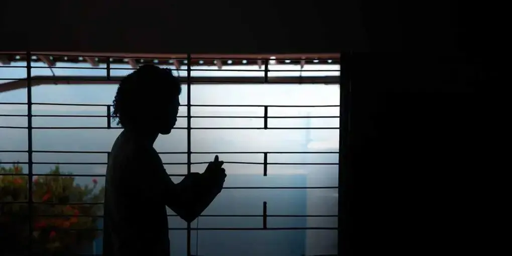 Silhueta de uma pessoa com cabelo cacheado em frente a uma janela com grades, iluminada pela luz externa, em ambiente interno escuro para matéria sobre absolvição de acusados de estupro de vulnerável e como proteger as meninas.
