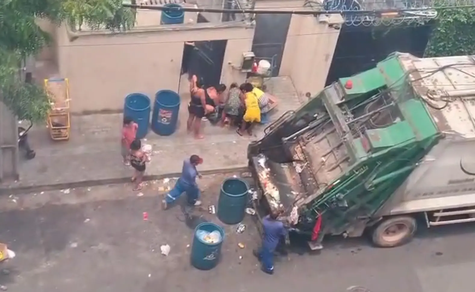 Vista aérea de um caminhão de lixo verde e branco coletando resíduos, com várias pessoas, incluindo crianças e dois coletores, interagindo com o lixo na rua.
