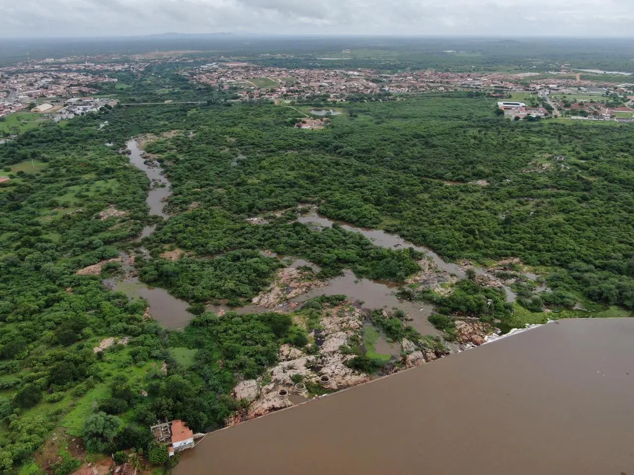 Vista aérea do açude do Batalhão com águas turvas, cercado por vegetação densa e a cidade de Crateús ao fundo sob um céu nublado.
