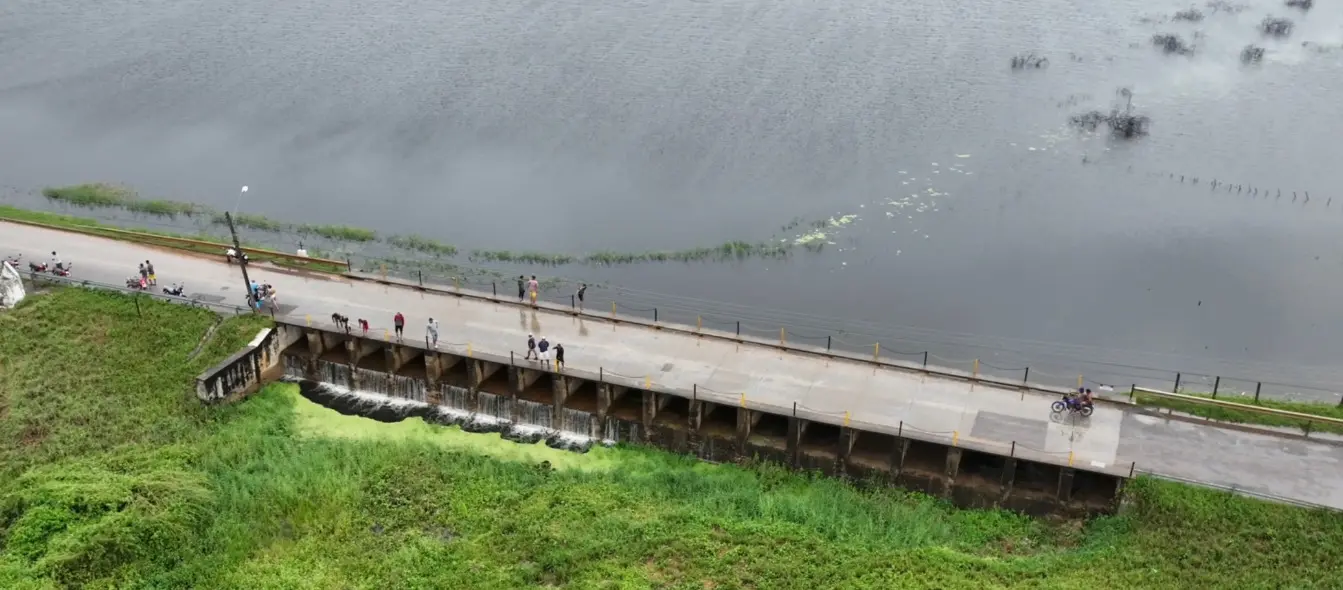 Vista aérea de uma ponte-barragem de concreto, no açude São José III, em Ipaporanga (CE), onde a água de um grande reservatório escoa por múltiplas comportas, formando pequenas quedas em meio a vegetação exuberante. Pessoas observam e motocicletas transitam na estrada superior.