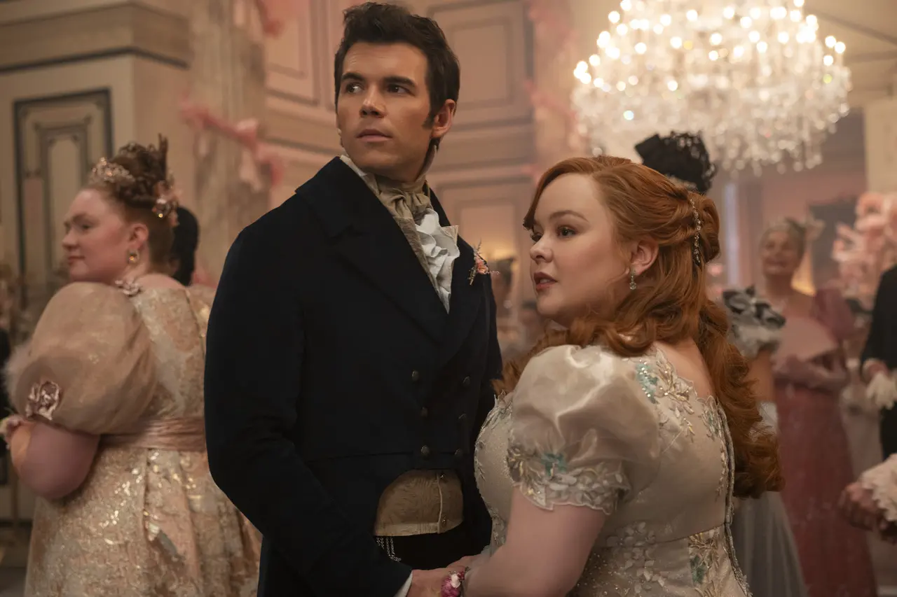 Cena de um baile de época em um grande salão decorado com tons suaves e detalhes florais. No centro, duas pessoas estão posicionadas frente a frente, segurando as mãos, vestindo trajes formais característicos do período regencial — uma com um casaco escuro com colete claro, outra com um vestido claro ornamentado. Ao fundo, outras pessoas circulam pelo salão, que é iluminado por um grande lustre de cristal pendurado no teto, e adornado com arranjos e laços rosados nas paredes.