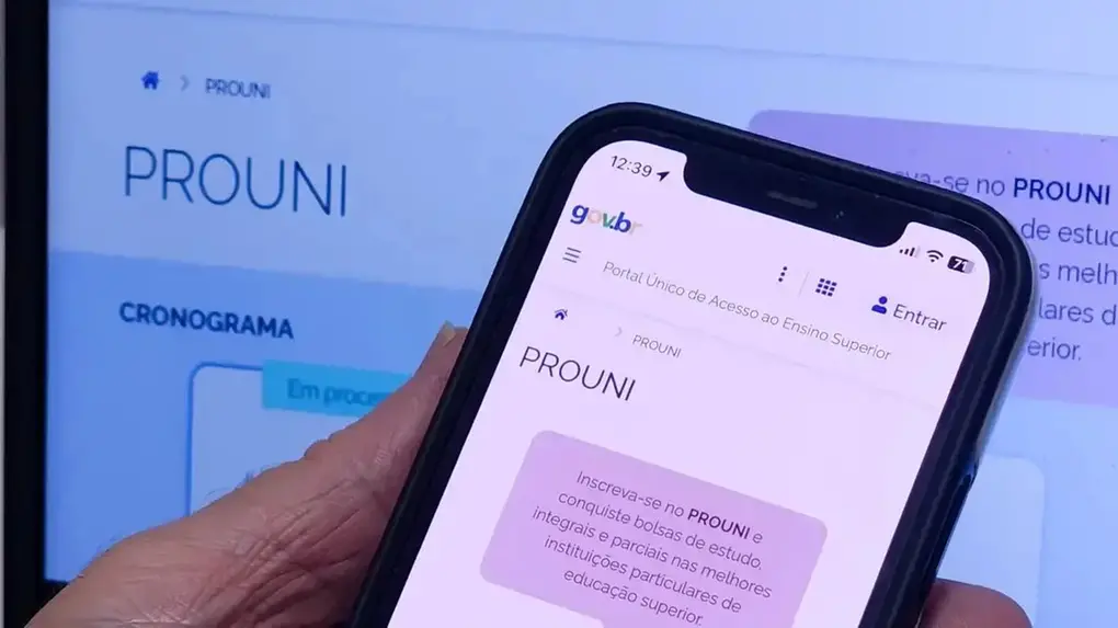 Uma mão segura um smartphone em primeiro plano, exibindo a página do Prouni no Portal Único de Acesso ao Ensino Superior, com o site correspondente visível ao fundo em um monitor de computador. Imagem usada em matéria sobre a segunda chamada do programa.