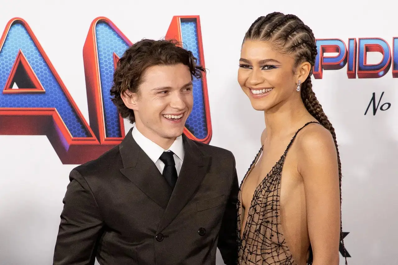 Os atores Tom Holland e Zendaya sorriem um para o outro em um tapete vermelho, com Holland vestindo um terno marrom escuro e Zendaya em um vestido de teia de aranha adornado com cristais. Foto usada em matéria sobre confirmação do casamento secreto dos dois.