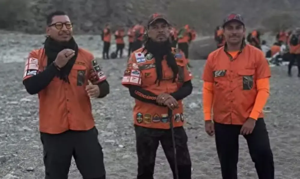 Três pessoas usando uniformes laranja com vários patches e brasões estão em uma área externa de terreno rochoso. As três estão posicionadas em pé na parte frontal da imagem, enquanto um grupo maior, também uniformizado, aparece ao fundo, distribuído pelo mesmo ambiente. O cenário é aberto, com chão coberto por pedras e formações rochosas ao redor.