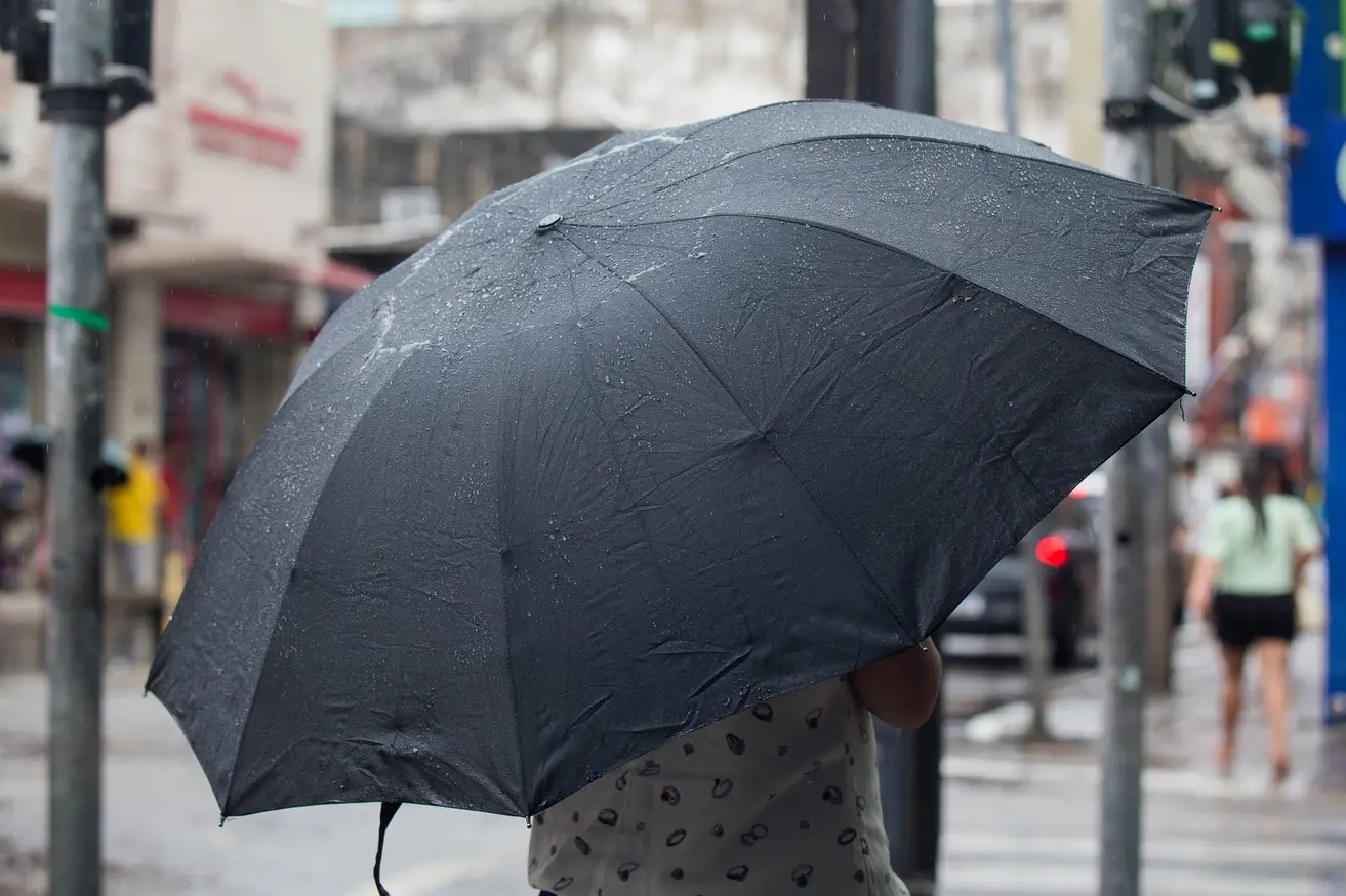 Pessoa segurando um guarda-chuva preto com gotas de chuva visíveis em uma rua movimentada da cidade.