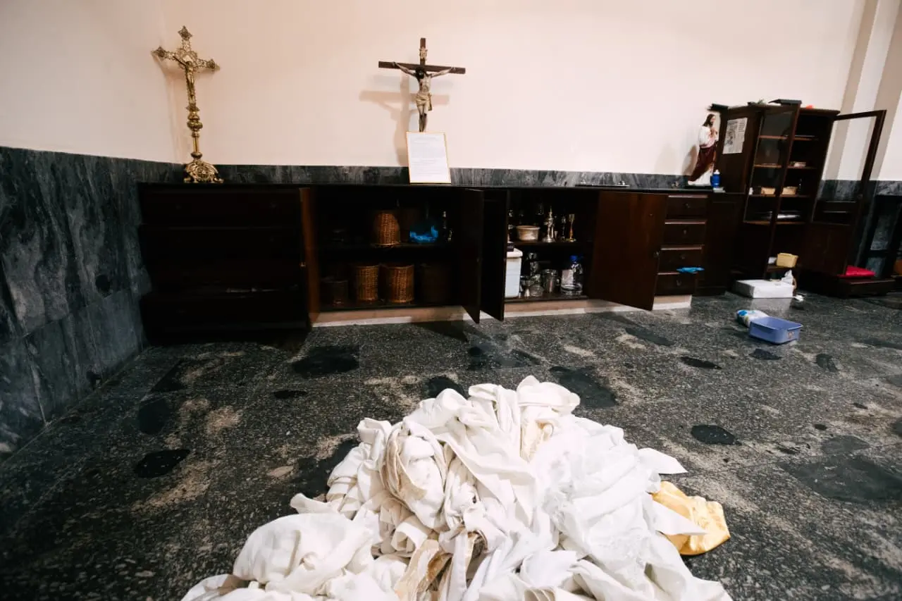 Interior de uma sacristia ou quarto de armazenamento de igreja com armários abertos revelando itens religiosos, dois crucifixos na parede, e uma grande pilha de tecidos brancos no chão de granito escuro com manchas escuras.