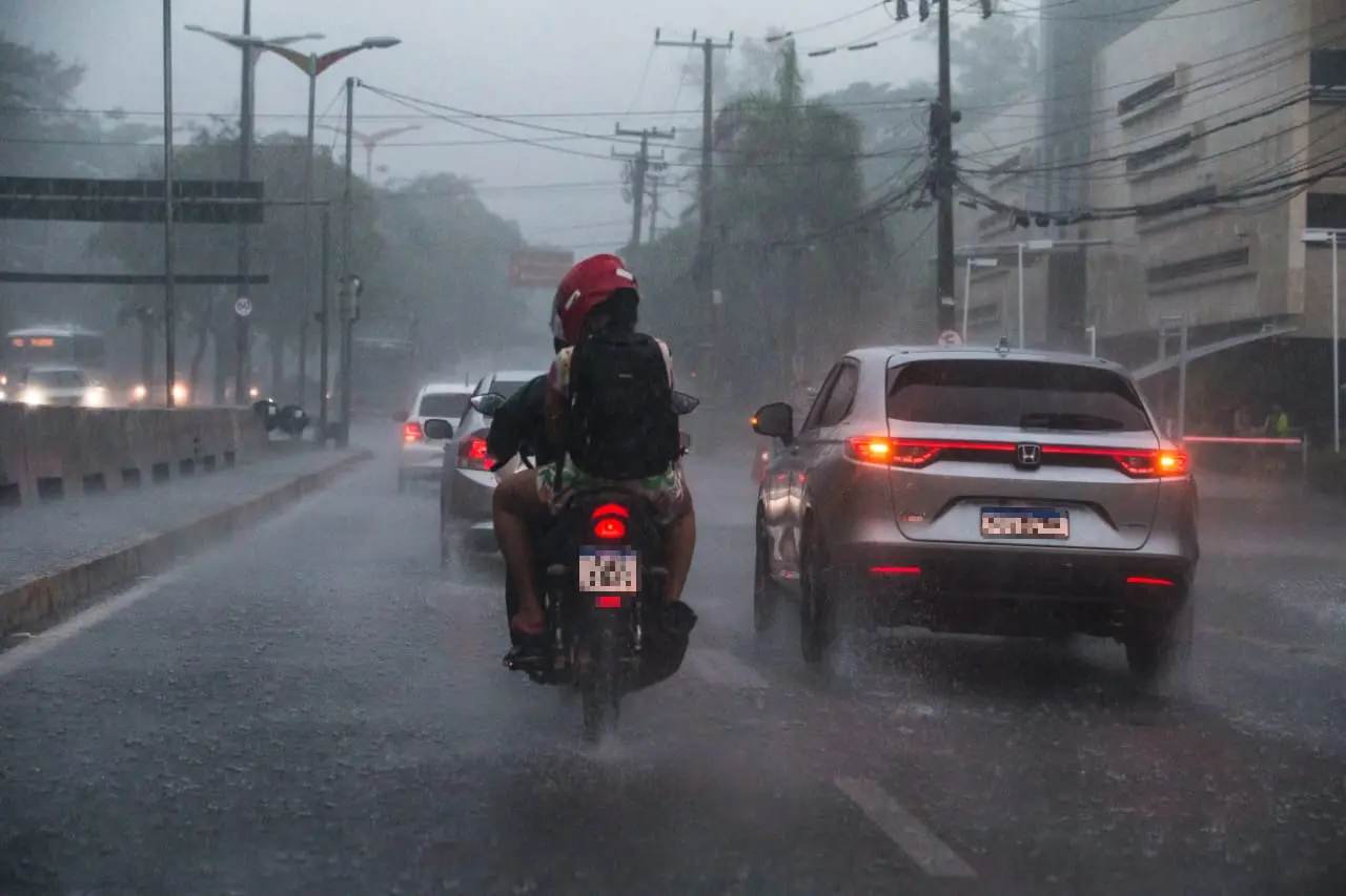 Motociclista e carros trafegando por rua urbana sob chuva forte, com asfalto molhado e respingos visíveis.
