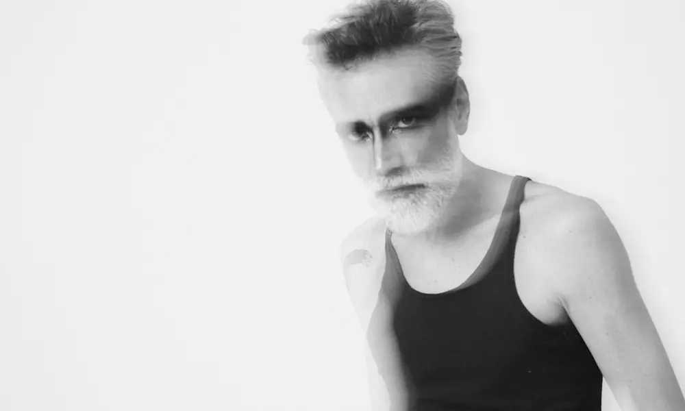 O músico Catatau, da banda Cidadão Instigado, encara a câmera em um retrato em preto e branco com efeito de sobreposição e leve desfoque. Ele usa uma regata escura, exibe uma barba branca destacada e uma maquiagem marcante que atravessa a linha dos olhos, criando uma composição artística e intensa.