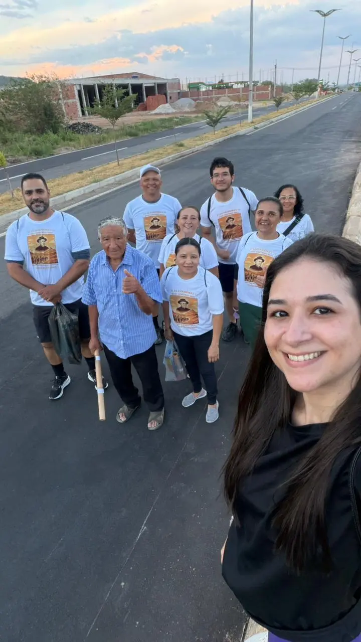 Na imagem, uma selfie ao ar livre em plano médio captura um grupo de nove pessoas reunidas em uma estrada asfaltada e larga, sob um céu de fim de tarde. Em primeiro plano, à direita, uma mulher jovem com longos cabelos escuros sorri para a câmera. Atrás dela, o grupo é liderado por um senhor idoso de camisa social azul clara listrada, que faz um sinal de positivo com o polegar. As outras sete pessoas (quatro mulheres e três homens de idades variadas) vestem camisetas brancas idênticas, estampadas com a imagem de uma figura religiosa e dizeres que sugerem uma caminhada ou evento comemorativo. O cenário ao fundo mostra o canteiro central de uma avenida, algumas árvores jovens e construções em alvenaria sob um céu com tons suaves de laranja e azul.