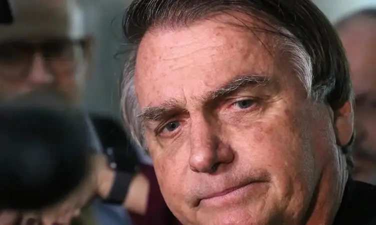Retrato em close do ex-presidente Jair Bolsonaro, apresentando uma expressão séria e olhar direcionado para baixo. Ele aparece em primeiro plano com iluminação lateral, enquanto figuras desfocadas e equipamentos de imprensa compõem o fundo da imagem.