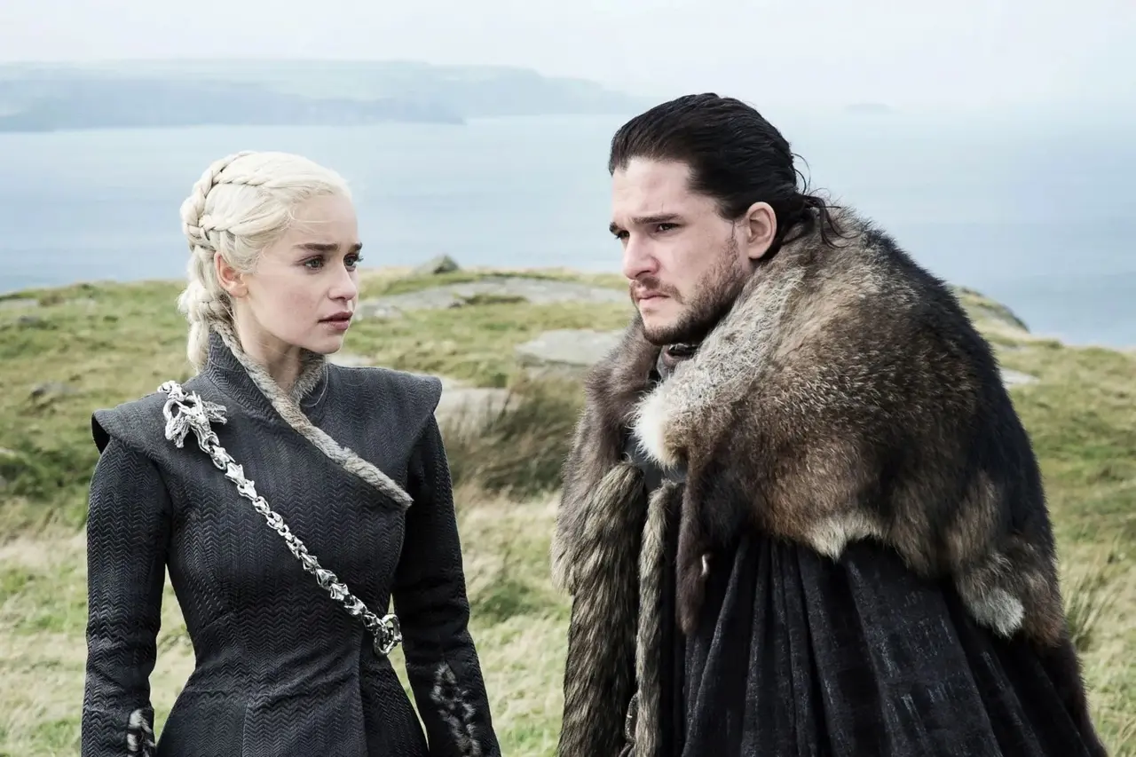 Personagens de 'Game of Thrones', Daenerys Targaryen e Jon Snow conversam, ambos com expressões sérias e vestindo trajes de inverno pesados e escuros. Daenerys destaca-se com seu cabelo platinado trançado e uma corrente de dragão, enquanto Jon usa uma robusta capa de pele sob um céu nublado.