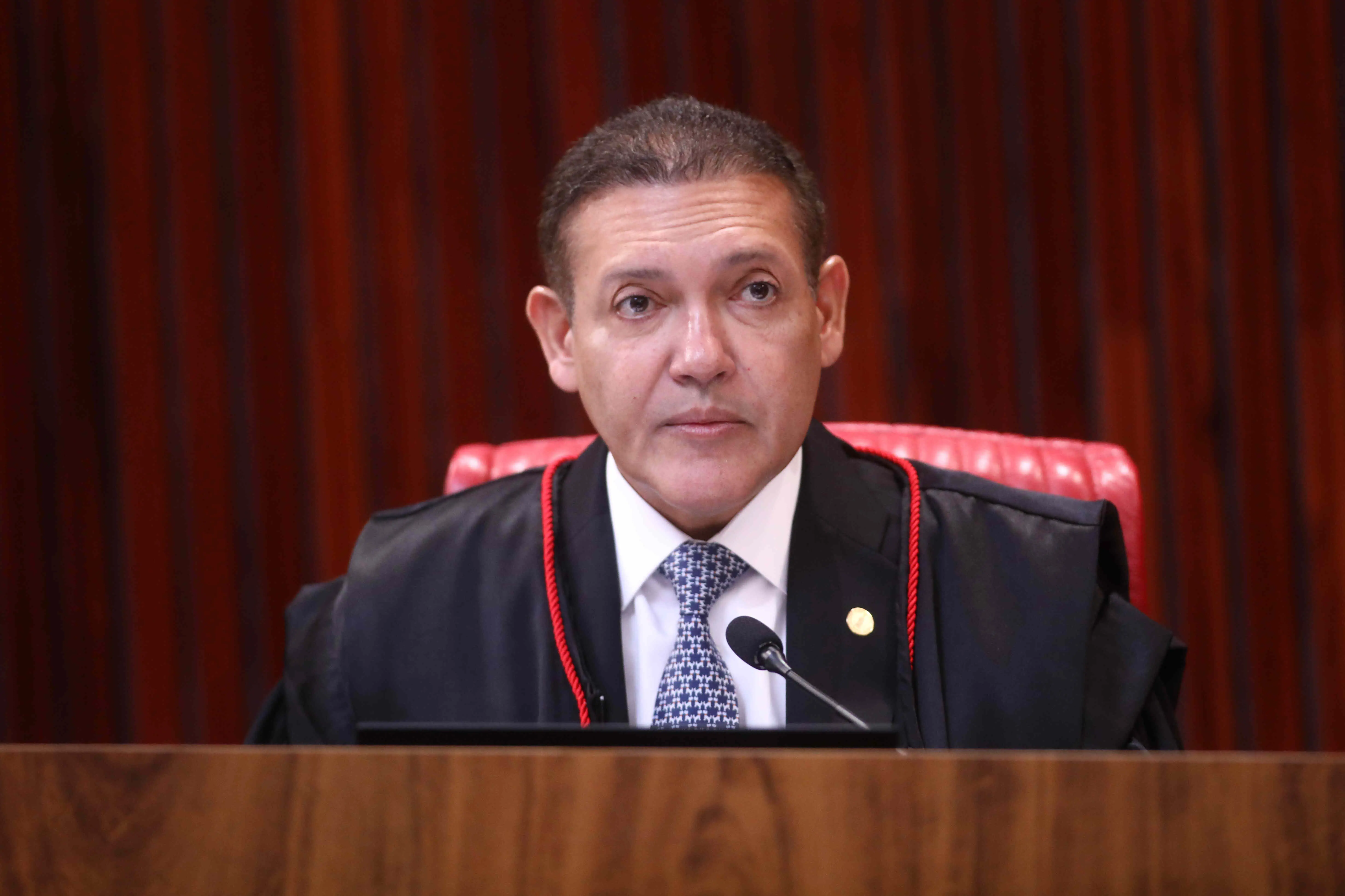 Homem de meia-idade, com cabelos curtos e escuros, sentado atrás de uma bancada de madeira, usando toga preta com detalhes em vermelho sobre terno, camisa branca e gravata estampada. Trata-se de Nunes Marques. Ele está em frente a um microfone, com expressão séria, e ao fundo há uma parede com painéis de madeira em tom avermelhado e uma cadeira estofada vermelha.